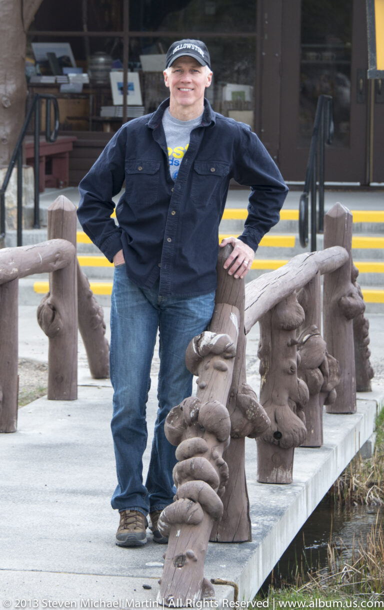 A visitor leans on a rustic log railing beside a lodge doorway and steps.