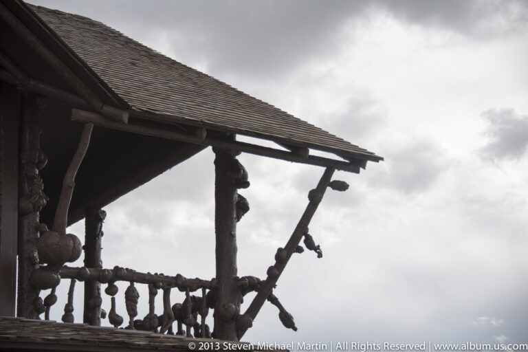 A shingled roof overhang rests on heavy timber brackets above a rustic log railing.