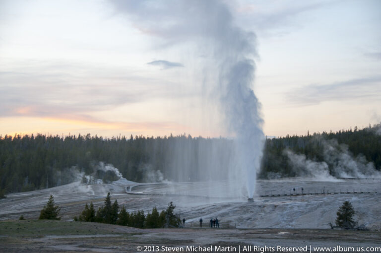 A geyser plume rises above a forested basin and boardwalk at sunset.