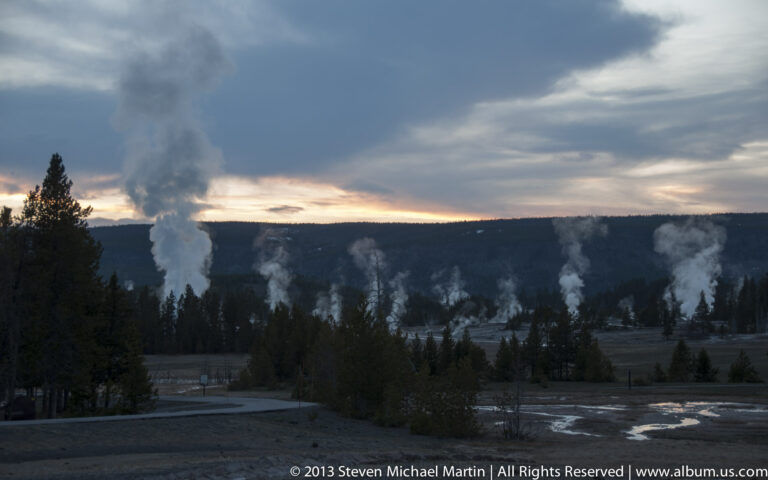 Multiple steam plumes rise from a wooded geothermal basin beneath a dim evening sky.