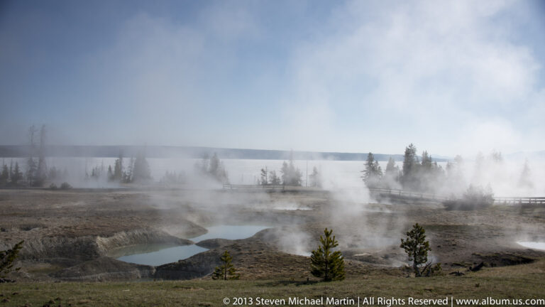 Steam drifts over small geothermal pools and pale ground beside a boardwalk.