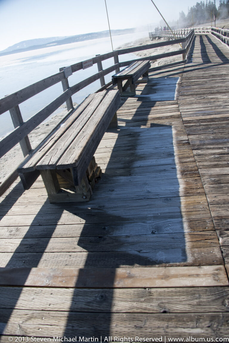 A wooden boardwalk and railings cast long shadows across a geothermal area.