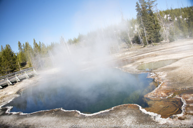 Steam drifts over a blue hot spring edged with orange deposits and a boardwalk.