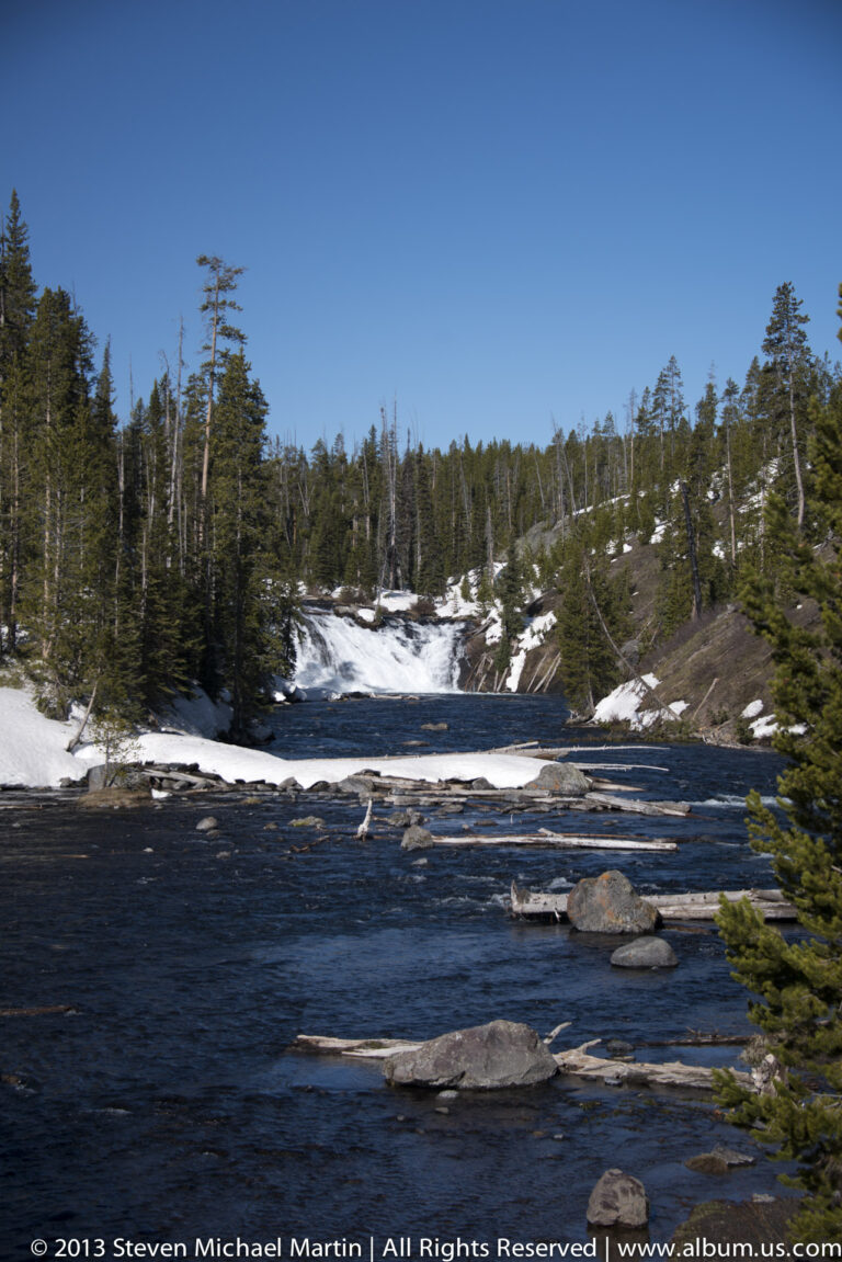 A dark river flows past rocks and a snow-lined bank beneath conifers.
