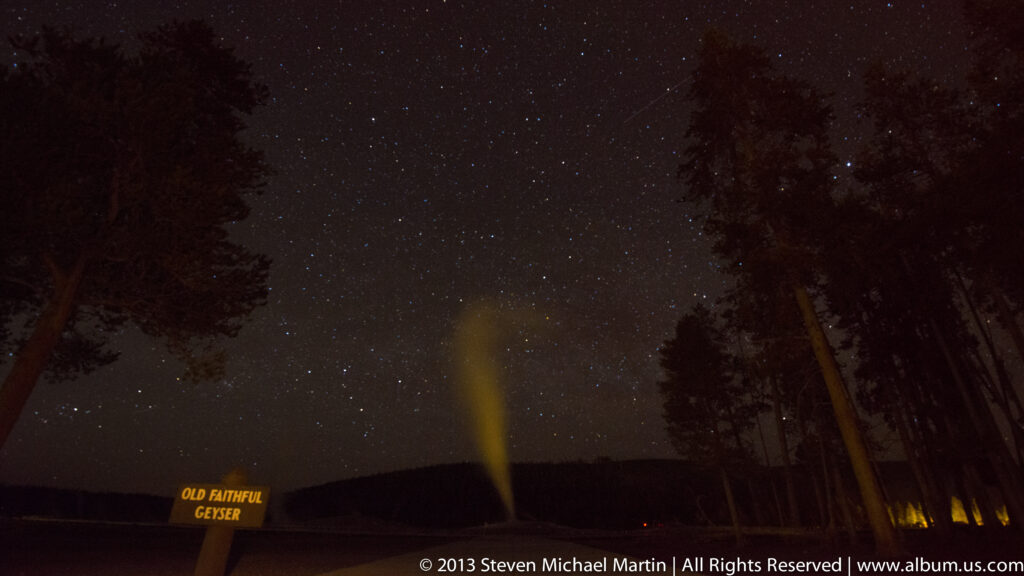 Old Faithful sends a yellow-lit plume into a starry sky, with dark trees and a small identifying sign in the foreground.
