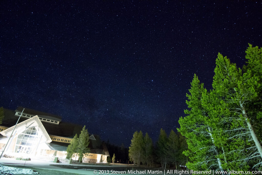 A glass-fronted, gabled lodge and evergreen trees sit beneath a broad, star-filled night sky.