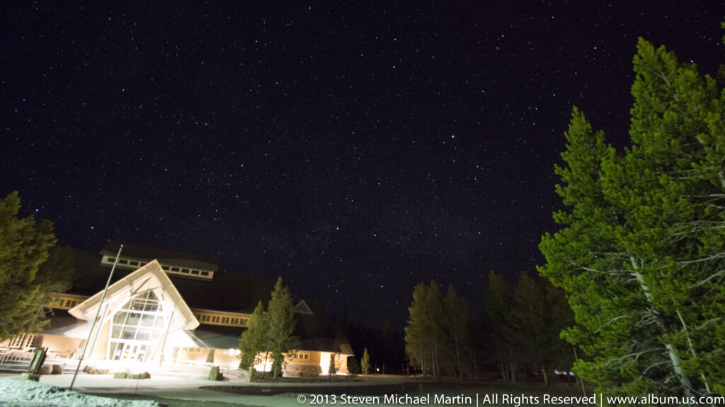 A brightly illuminated glass-gabled lodge stands below a starry sky, framed by a tall pine and smaller trees.