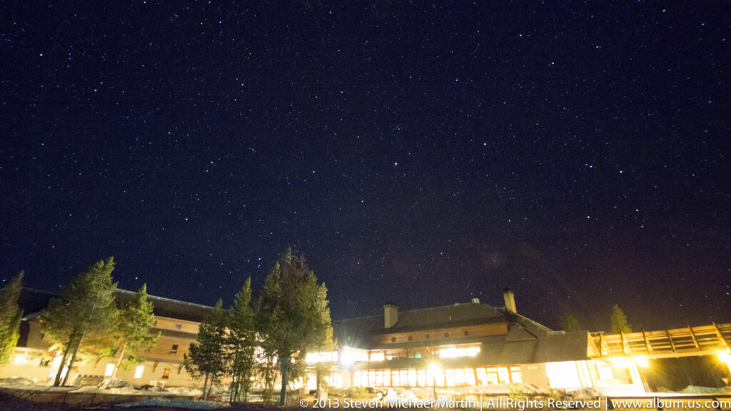 A long, brightly lit lodge stretches behind several trees beneath a dark blue sky dotted with stars.