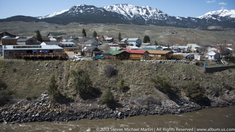 A riverside town sits below snow-covered mountains, viewed across a rocky bank.