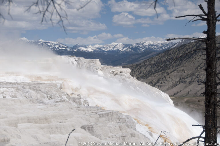 Pale travertine terraces descend toward a mountain valley, with steam rising at left.