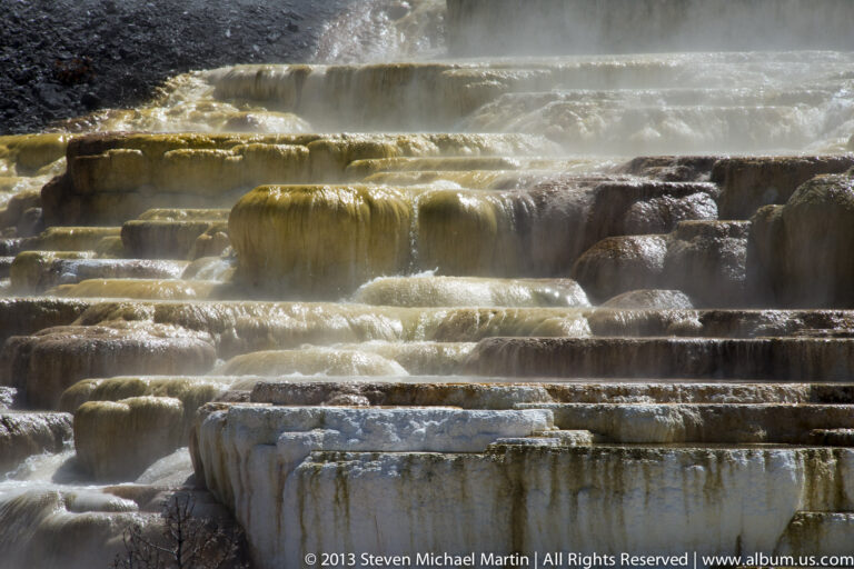 Golden water cascades over pale travertine steps as steam rises behind the pools.