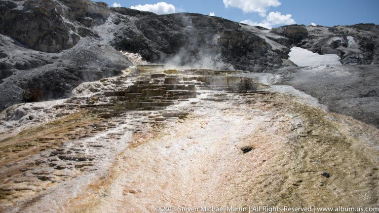Pale travertine terraces descend a dark hillside, with rusty bands and a small patch of snow above.