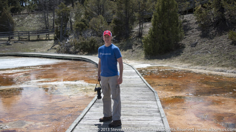 A visitor in a blue shirt and red cap stands on a boardwalk crossing an orange-stained thermal terrace.