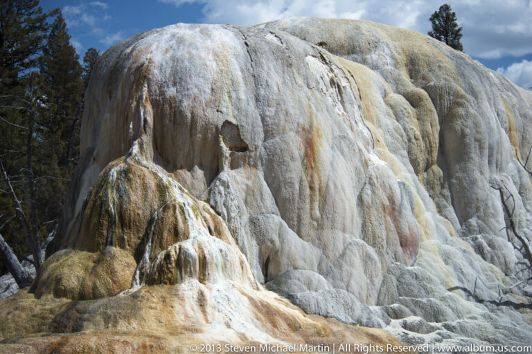 A pale travertine mound with orange streaks rises between conifers beneath a partly cloudy sky.