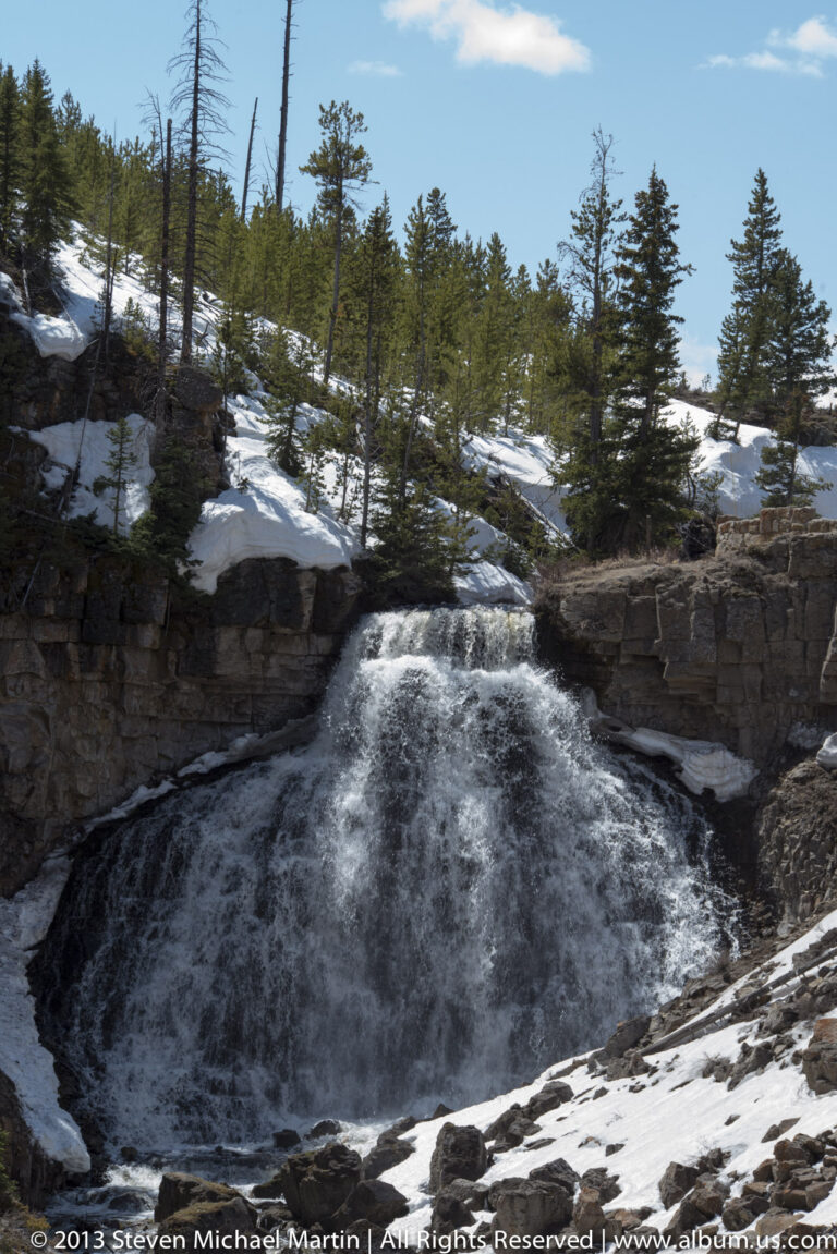 A waterfall pours over a dark ledge into a basin bordered by snow and evergreen trees.