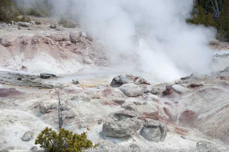 A dense steam plume rises over pale pink and gray mineral ground scattered with rocks.