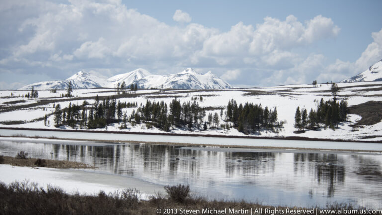 A dark river crosses a snow-covered valley beneath distant mountains and billowing clouds.