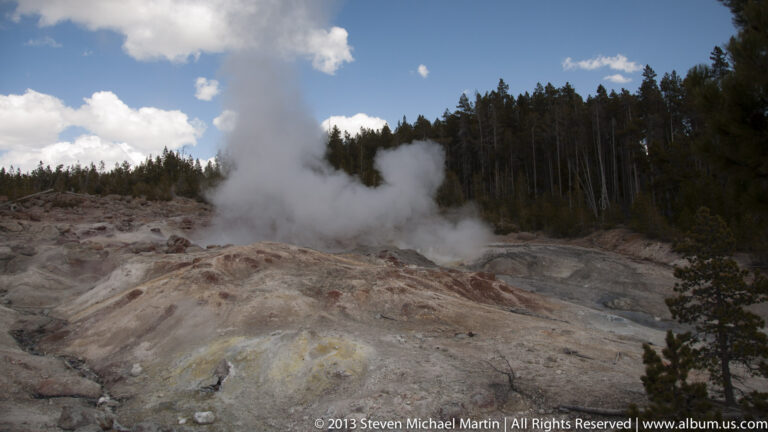 A large steam column rises from a pale geothermal mound in front of a dark forest.