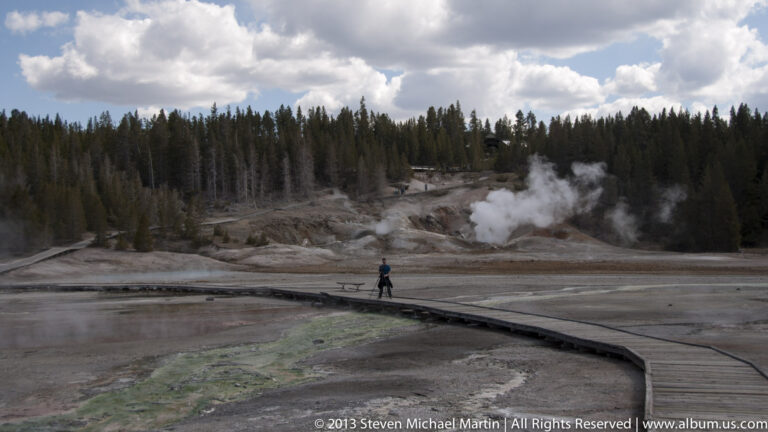 A lone visitor walks along a curving boardwalk through a pale basin with rising steam.