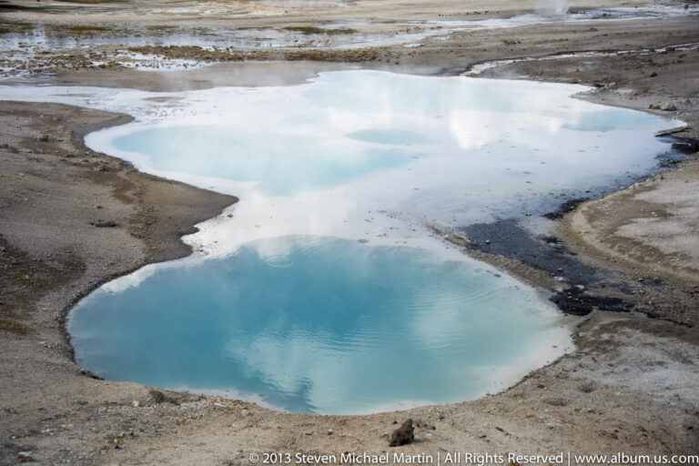 Two turquoise pools with pale mineral rims sit in a bare geothermal basin.