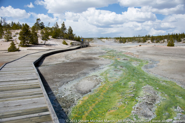 A curving boardwalk crosses a thermal basin beside vivid green and orange mineral mats.