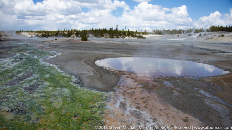 Shallow pools and green-orange runoff spread across a broad thermal flat beneath a cloudy sky.