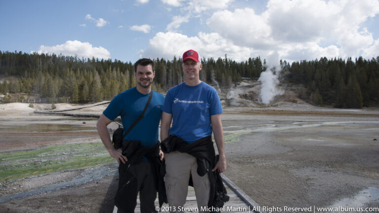 Two men pose on a boardwalk with a steaming geothermal basin and trees behind them.