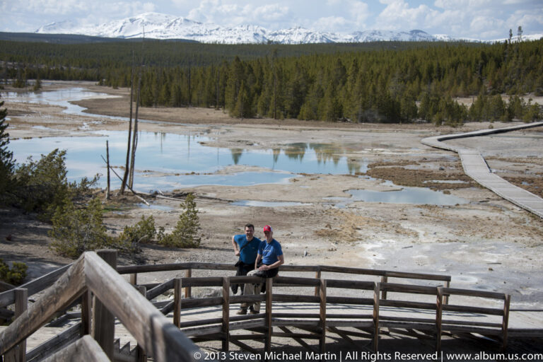 Two visitors sit on a wooden railing above a thermal basin crossed by a boardwalk.