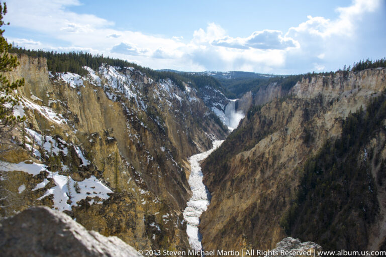 A distant waterfall and winding river sit within a broad, steep-sided canyon.
