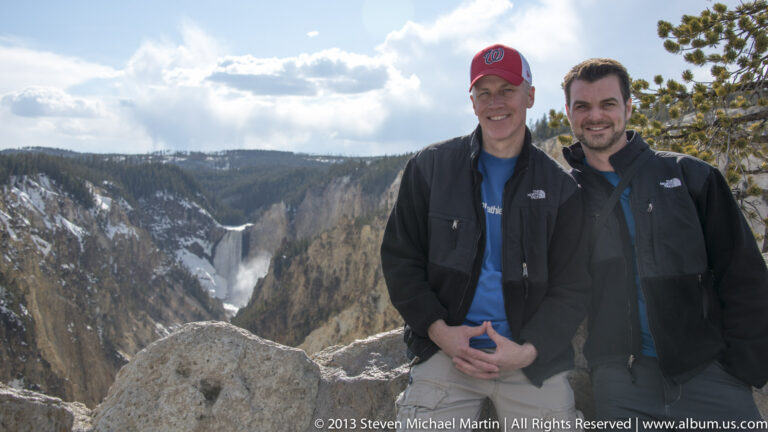 Two men stand at a rocky canyon overlook with a winding river and snow patches behind them.