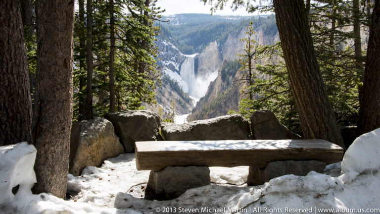 A wooden bench beneath conifers faces a snow-edged canyon and distant waterfall.