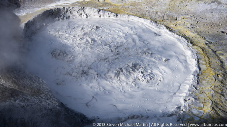 A pale thermal pool bubbles within a yellow-brown mineral rim as steam drifts across it.