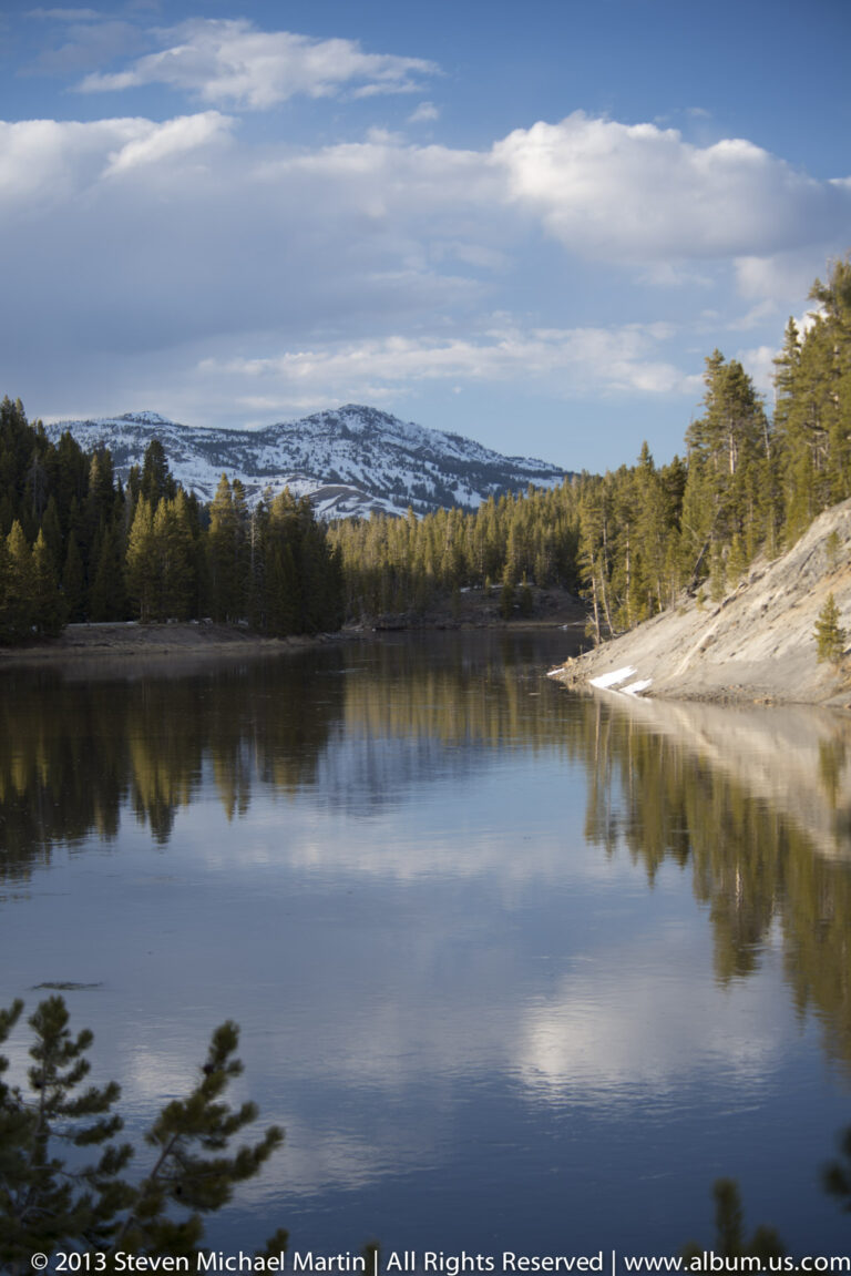 A still lake reflects evergreen trees and a snow-dusted ridge beneath a pale blue sky.