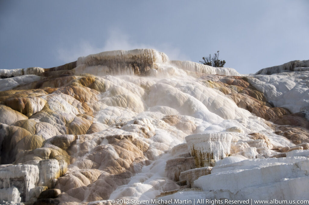 Broad cream and ochre travertine terraces descend a hillside as water flows through channels and steam rises above.