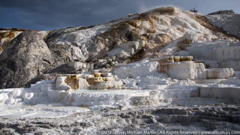 A dark boulder sits beside stepped white travertine terraces with small pools and orange mineral staining.