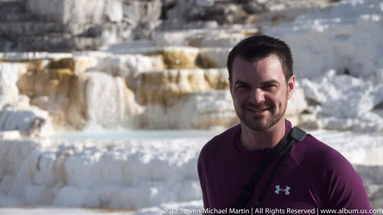 A man in a maroon shirt stands in the foreground, with pale travertine terraces softly blurred behind him.
