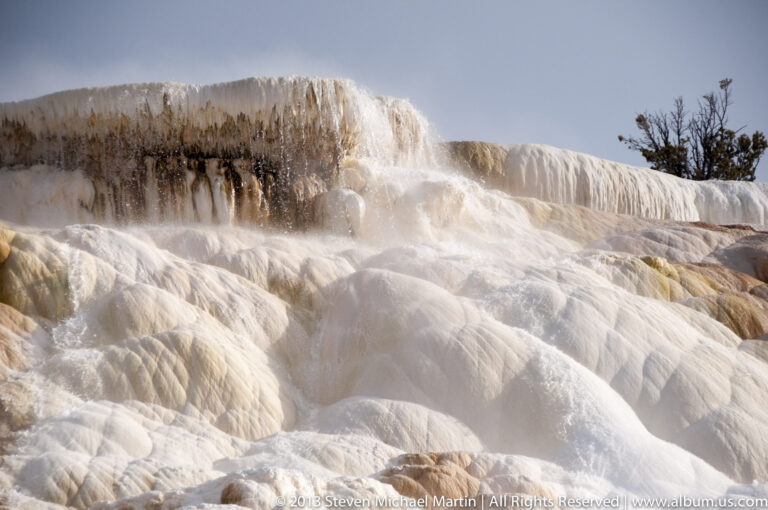 Steam rises from rounded cream and rust-colored travertine mounds as water runs over shallow ledges.