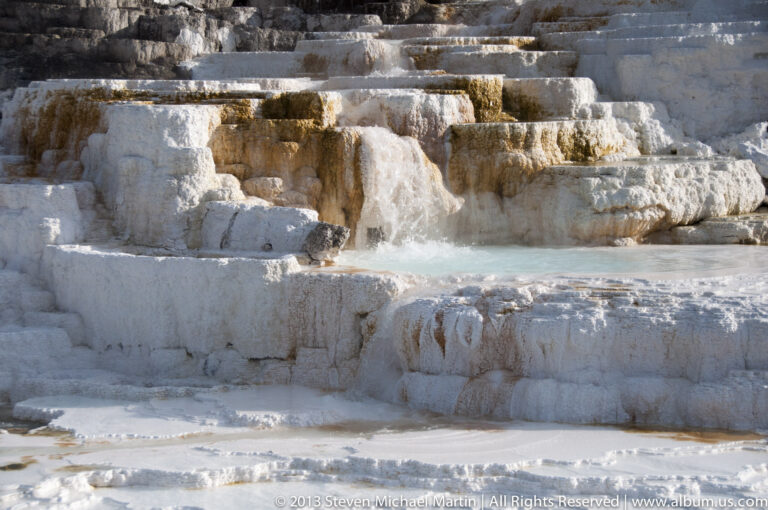 A pale blue thermal pool sits below white travertine shelves with scalloped edges and small cascades.