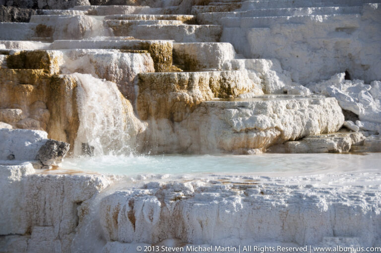 Water cascades over pale travertine steps into a turquoise pool beside a dark rock outcrop.