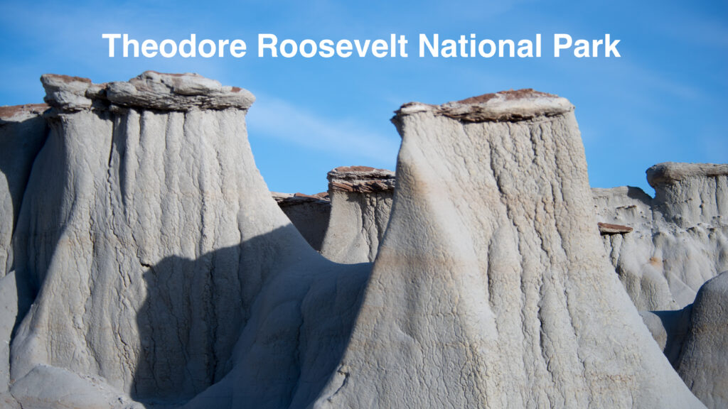 Pale, fluted claystone hoodoos with reddish caprock stand beneath a clear blue sky.