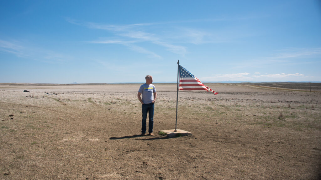 A visitor stands beside a United States flag on open, dry prairie beneath a wide blue sky.