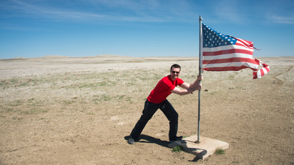 A man in a red shirt leans beside a flagpole as a United States flag billows over open prairie.