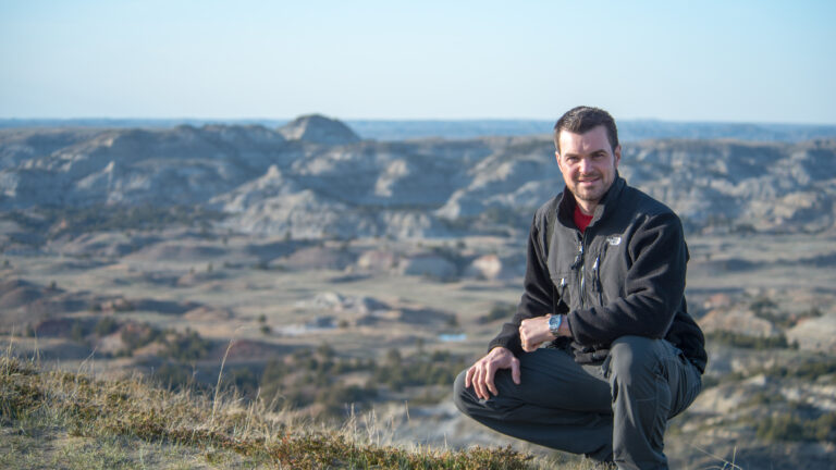 A man crouches in the foreground with softly focused badlands ridges behind him.
