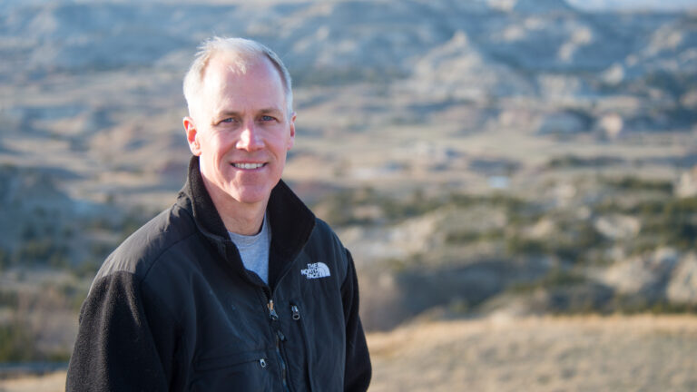 A man in a dark jacket smiles at the camera with blurred badlands behind him.