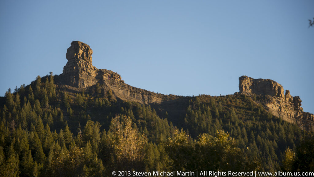 Two rock pinnacles stand along a forested ridge above autumn-colored trees beneath a clear blue sky.