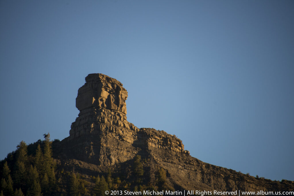 A broad-topped rock pinnacle rises above a dark tree line, its stepped ledges lit against a clear blue sky.