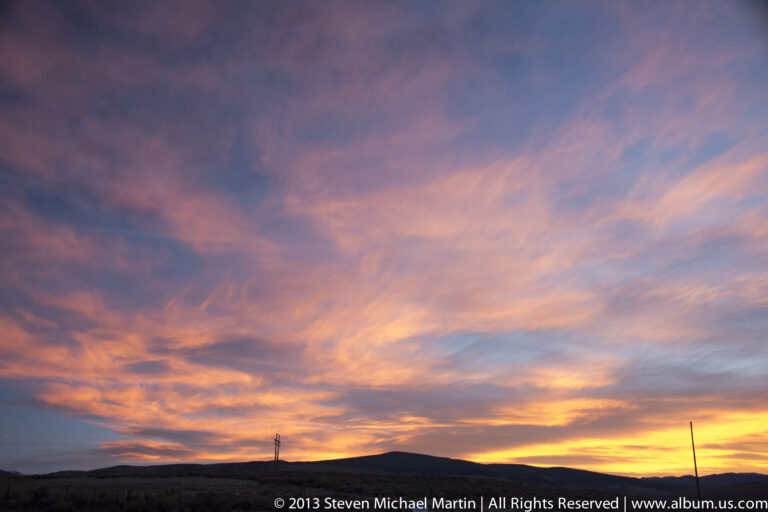 Pink and orange sunset clouds glow above a dark mountain ridge, with a slender pole at the right.
