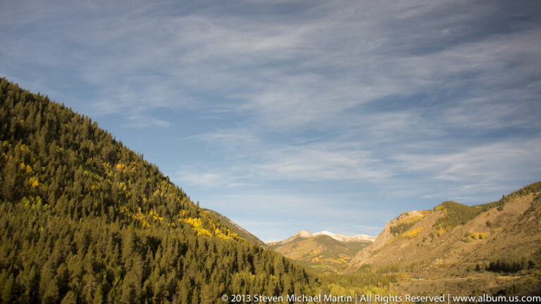 Golden aspens and dark conifers cover a mountain valley leading toward distant snow-dusted peaks.