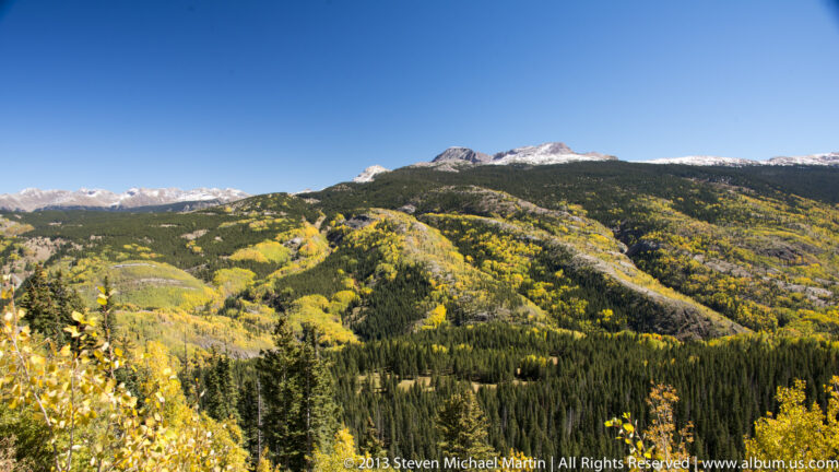 Golden aspens and evergreen forests spread across ridges beneath distant snow-dusted peaks.