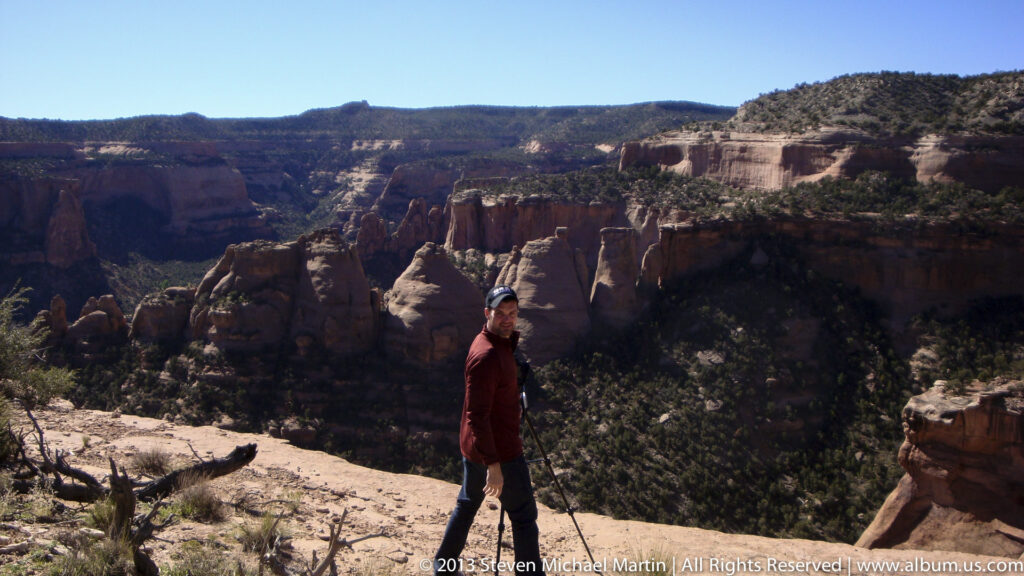 A visitor stands at a rocky overlook above a red-rock canyon with rounded formations and scrub-covered slopes.